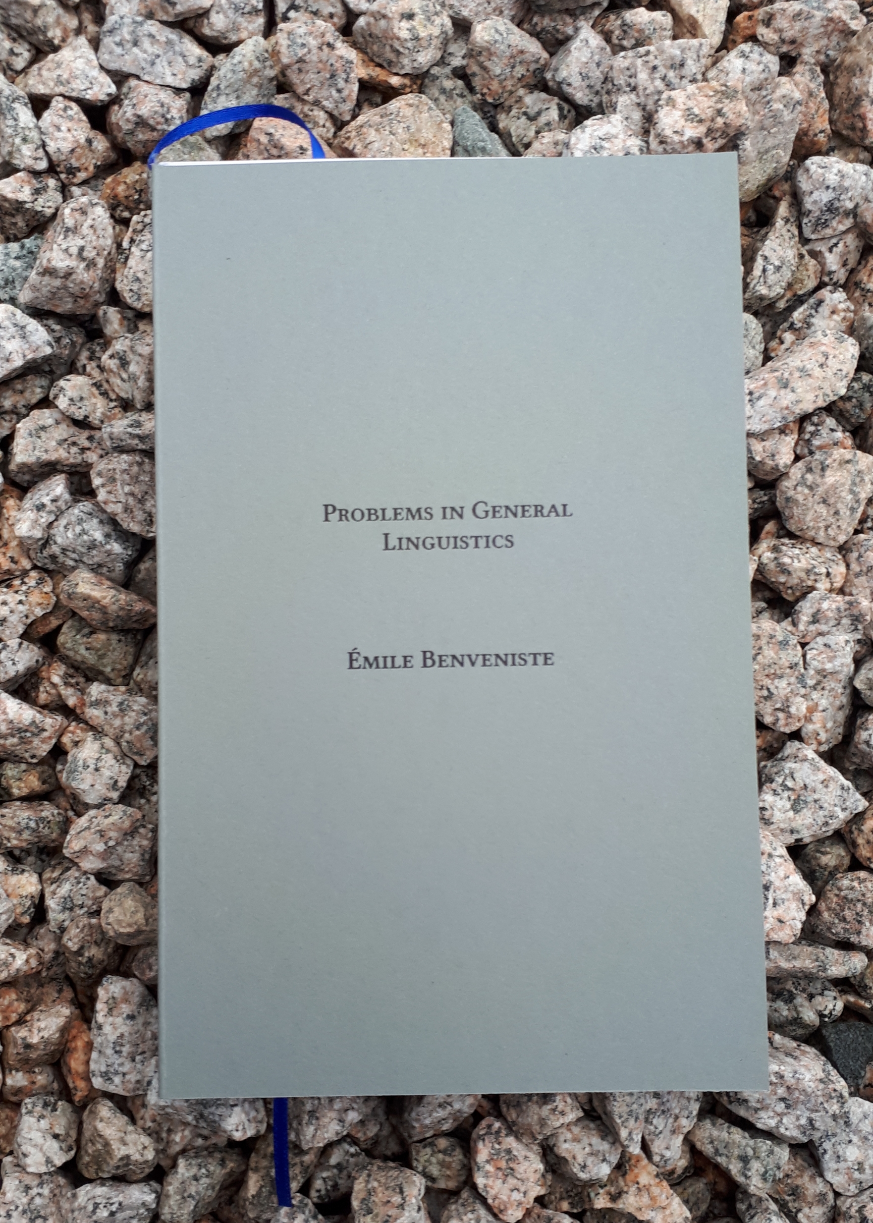 A book with a slate gray cover and blue marker ribbon lies on a bed of sharp gravel. Text on the cover reads "Problems in General Linguistics, Emile Benveniste"