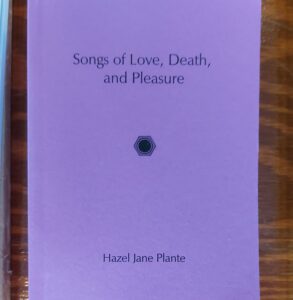 A book with a vibrant purple cover, displayed against a dark wooden tabletop. The title is Songs of Love, Death, and Pleasure by Hazel Jane Plante. Also on the cover is a drawing of a 1/4" audio jack, the kind you would plug an electric guitar into.